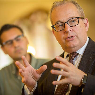 Motioning with his hands, Steve DeWeerth wears glasses and a suit and tie while speaking while an onlooker is seen blurred behind him.