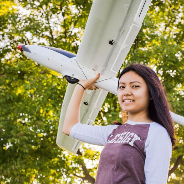 A female student wearing a baseball-style brown Lehigh shirt smiles while holding a large white airplane model above her head with one hand.