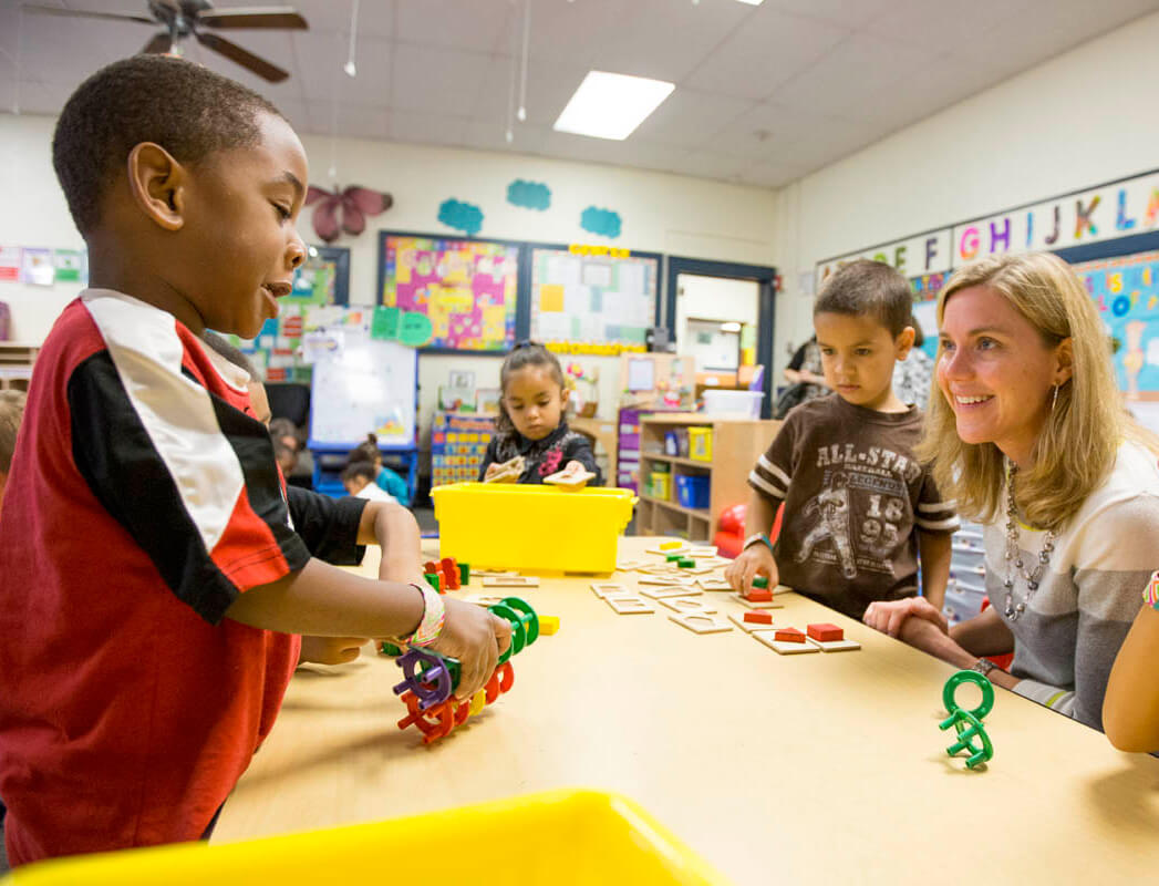 Teacher sits at a table with early elementary aged students, smiling as they work with colorful puzzles and building games