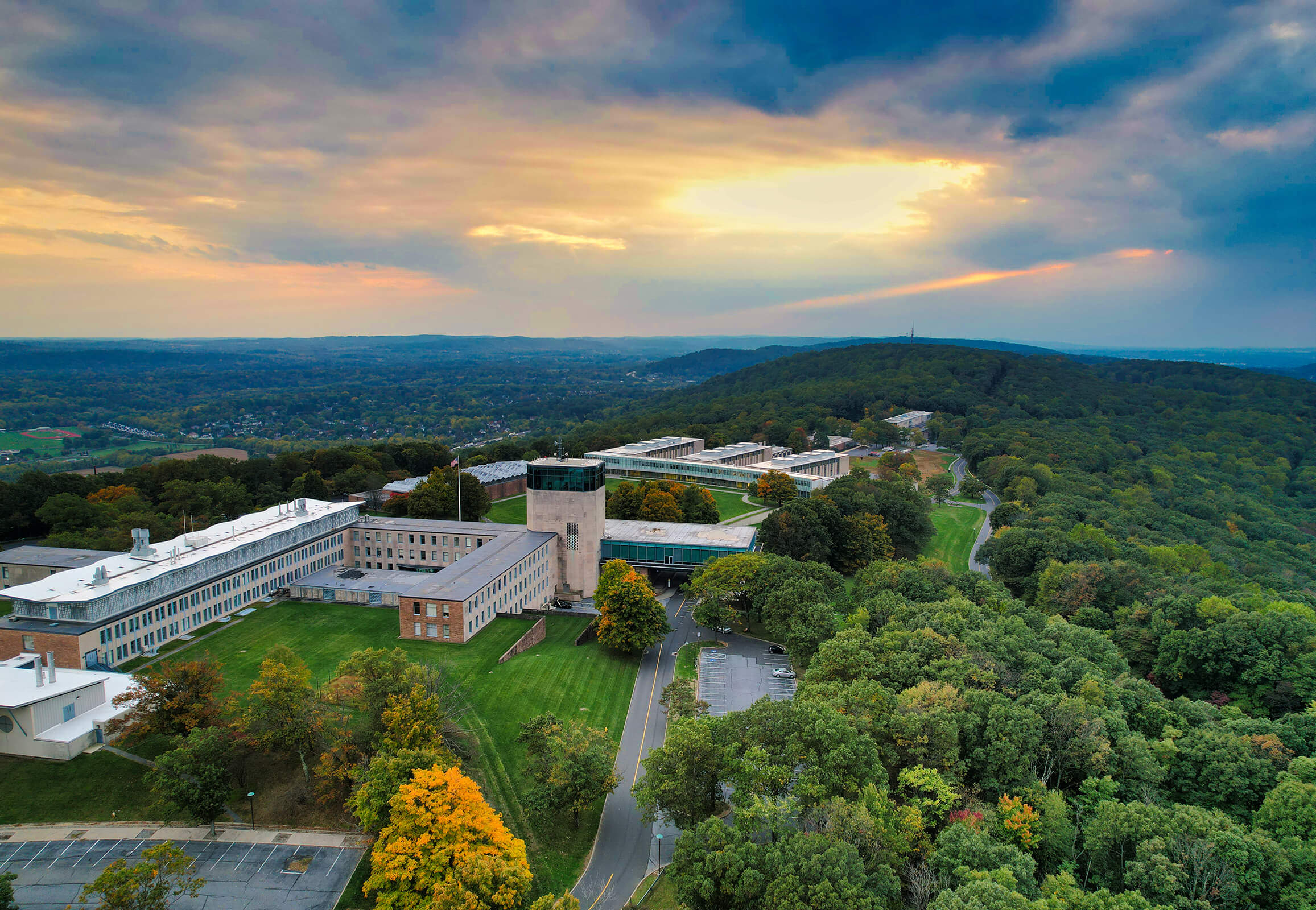 A aerial view of Mountaintop campus, showing the buildings surrounded by the green landscape under a twilight sun peaking through clouds.