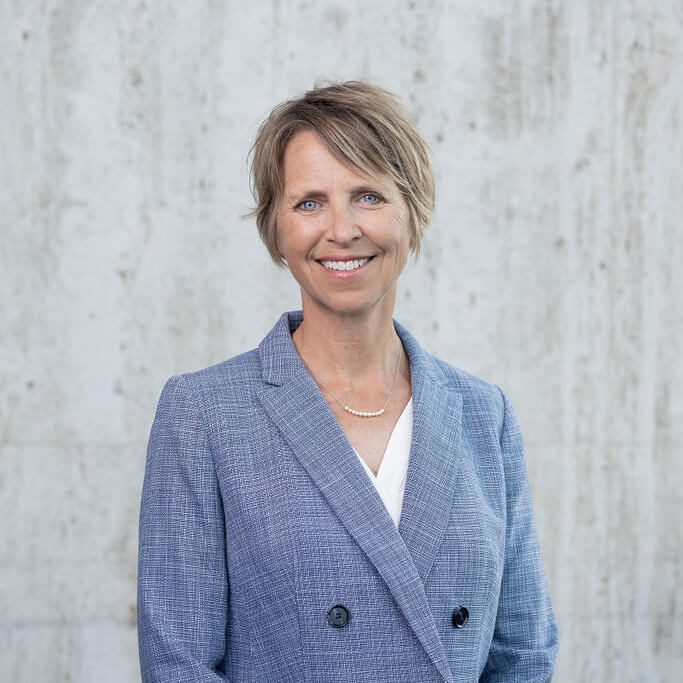 Woman with short blonde hair poses in front of a concrete wall, wearing a light blue double breasted blazer and a smile.