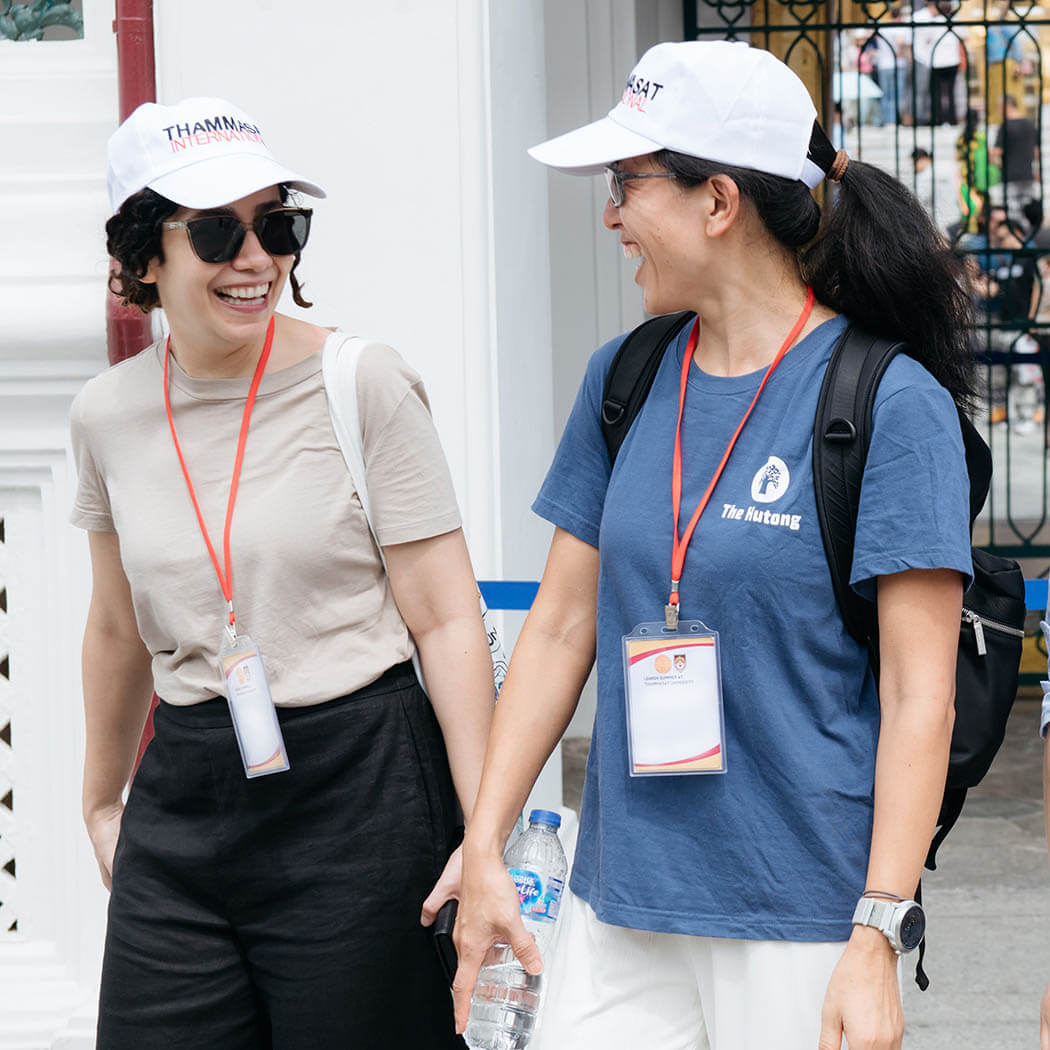 Two students wearing white Thomasset International hats and lanyards, smiling and laughing while looking at each other. 