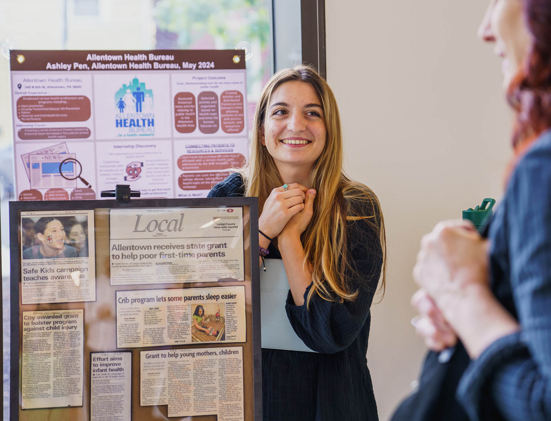 Student stands beside her presentations at the College of Health symposium with hands clasped and smiling as she presents
