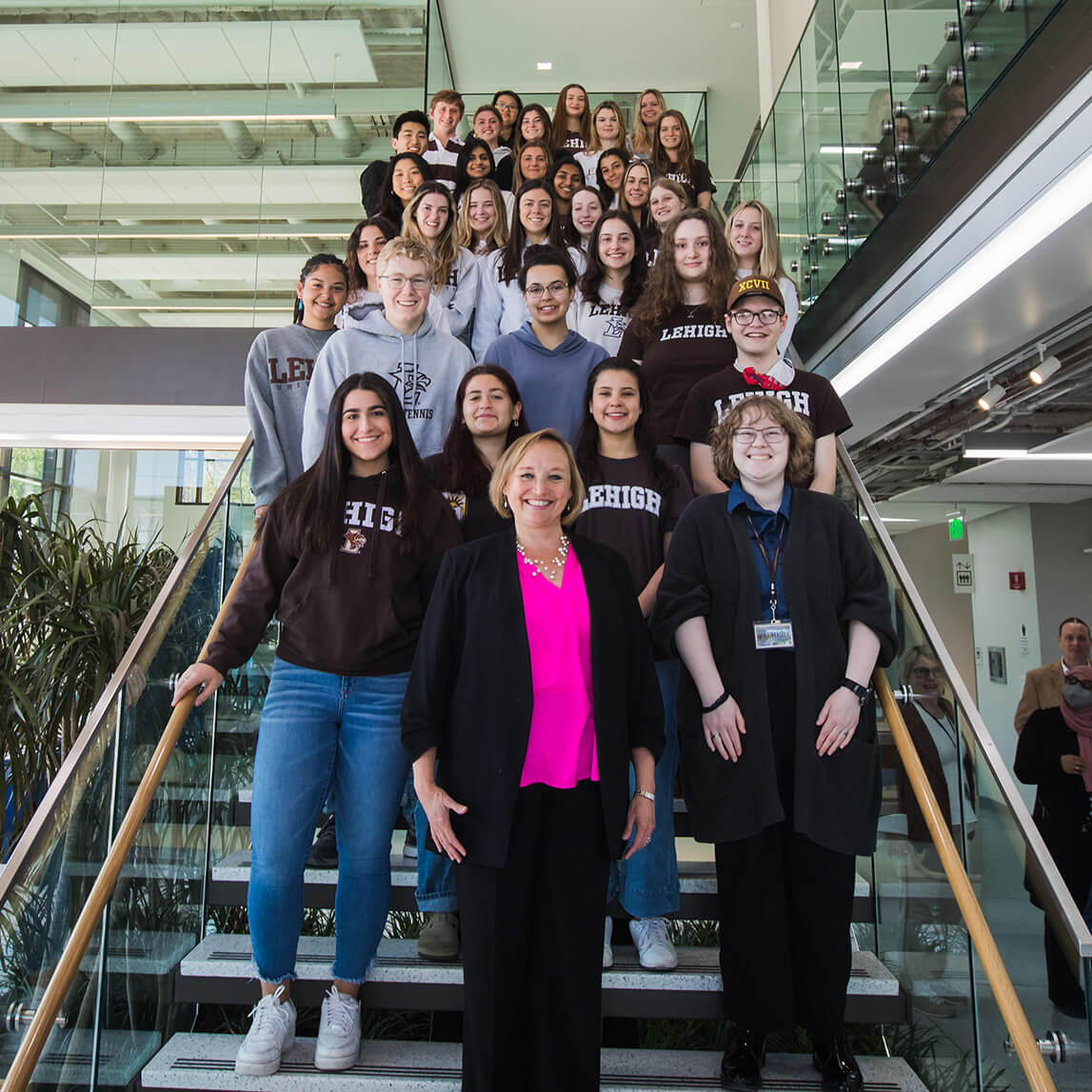 Dean Dolan poses on stairs in the Health, Science, and Technology building with a large group of college of health students standing on the stairs behind her