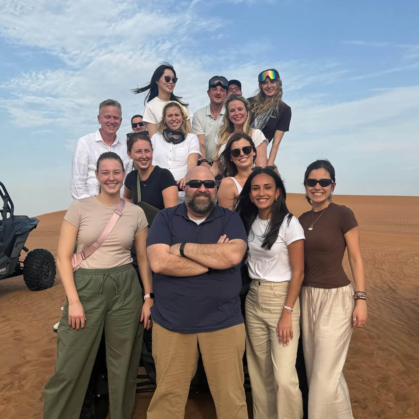 14 people pose in on a vast, sandy terrain, piled atop a dune buggy with another one shown off frame.