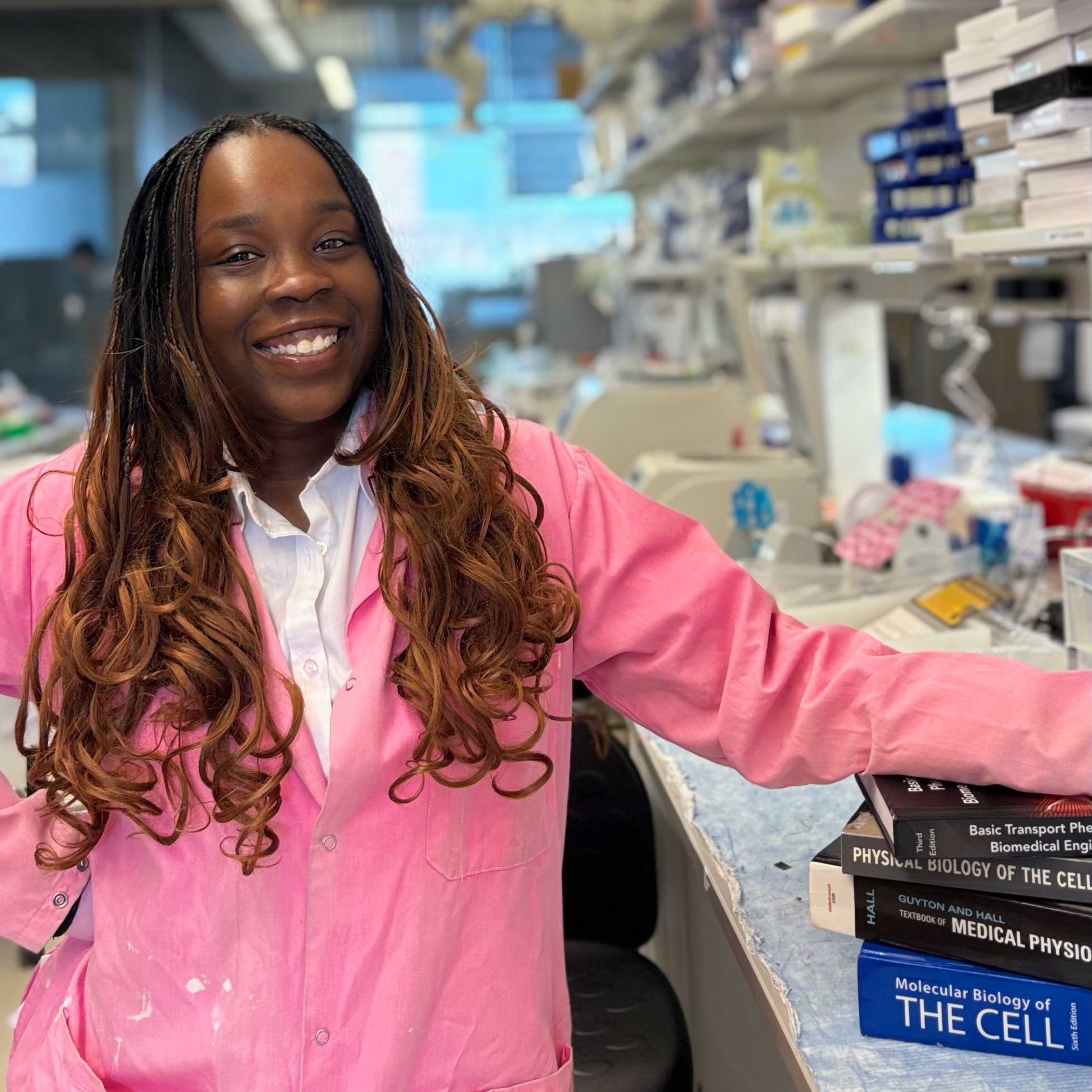 Aisha wears a bright pink lab coat with a white top underneath. She stands in a lab; her arms rests on biology research books.
