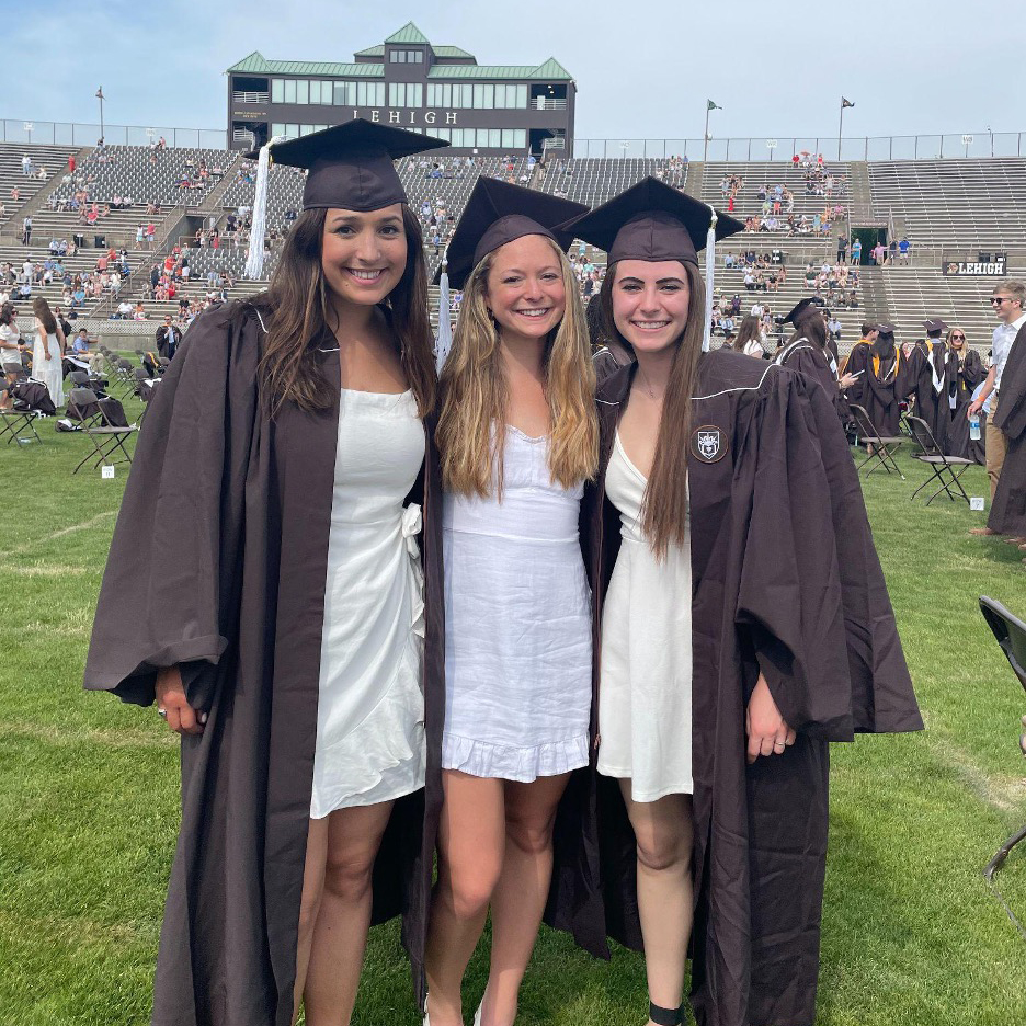 Ali, Julia, and their friend Allie stand together on the graduation field in their caps and gowns. They wear white dresses.