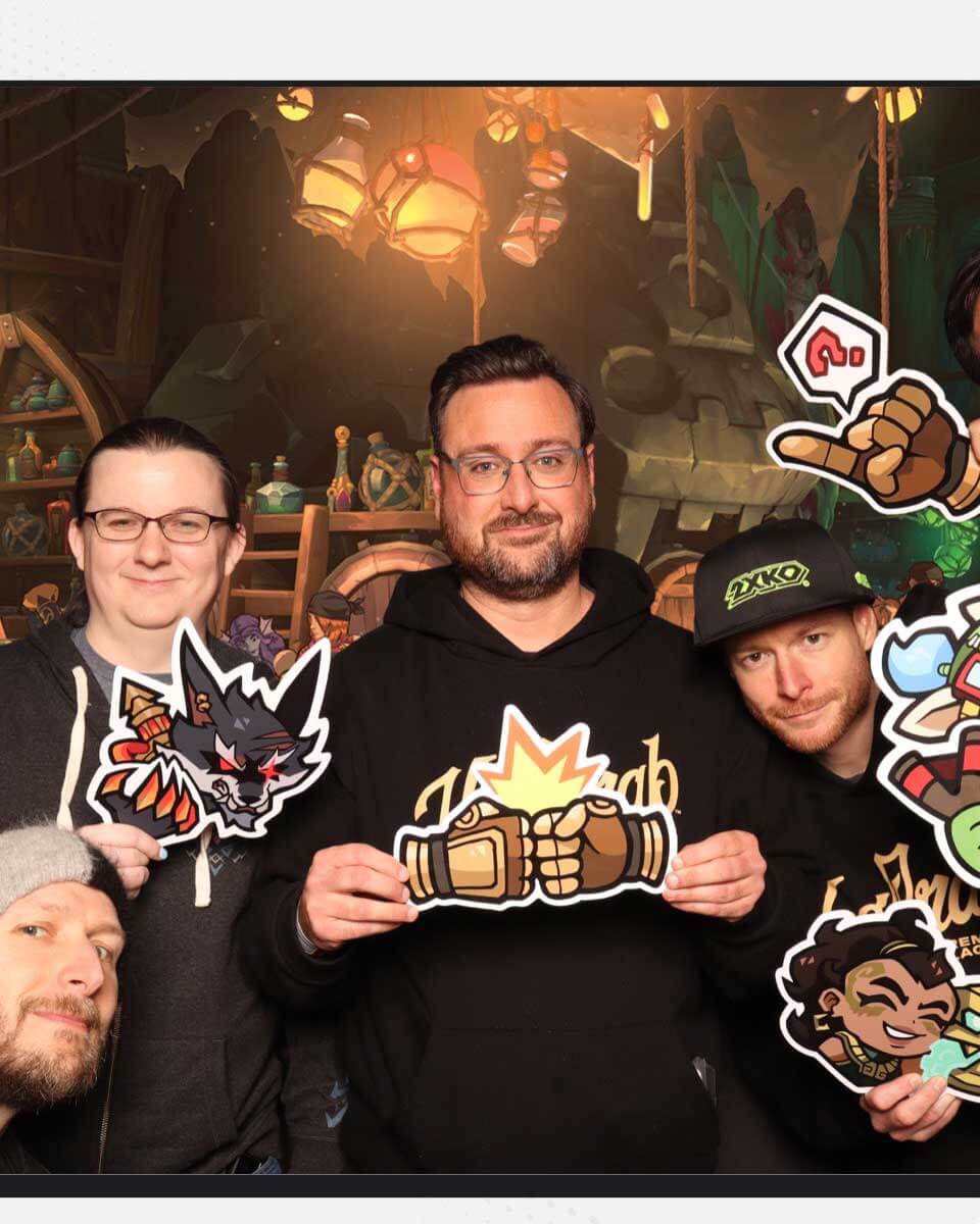 Andrew and his coworkers smile while holding cutouts with motifs and characters from 2XKO; Andrew's shows two fists mid-bump. 