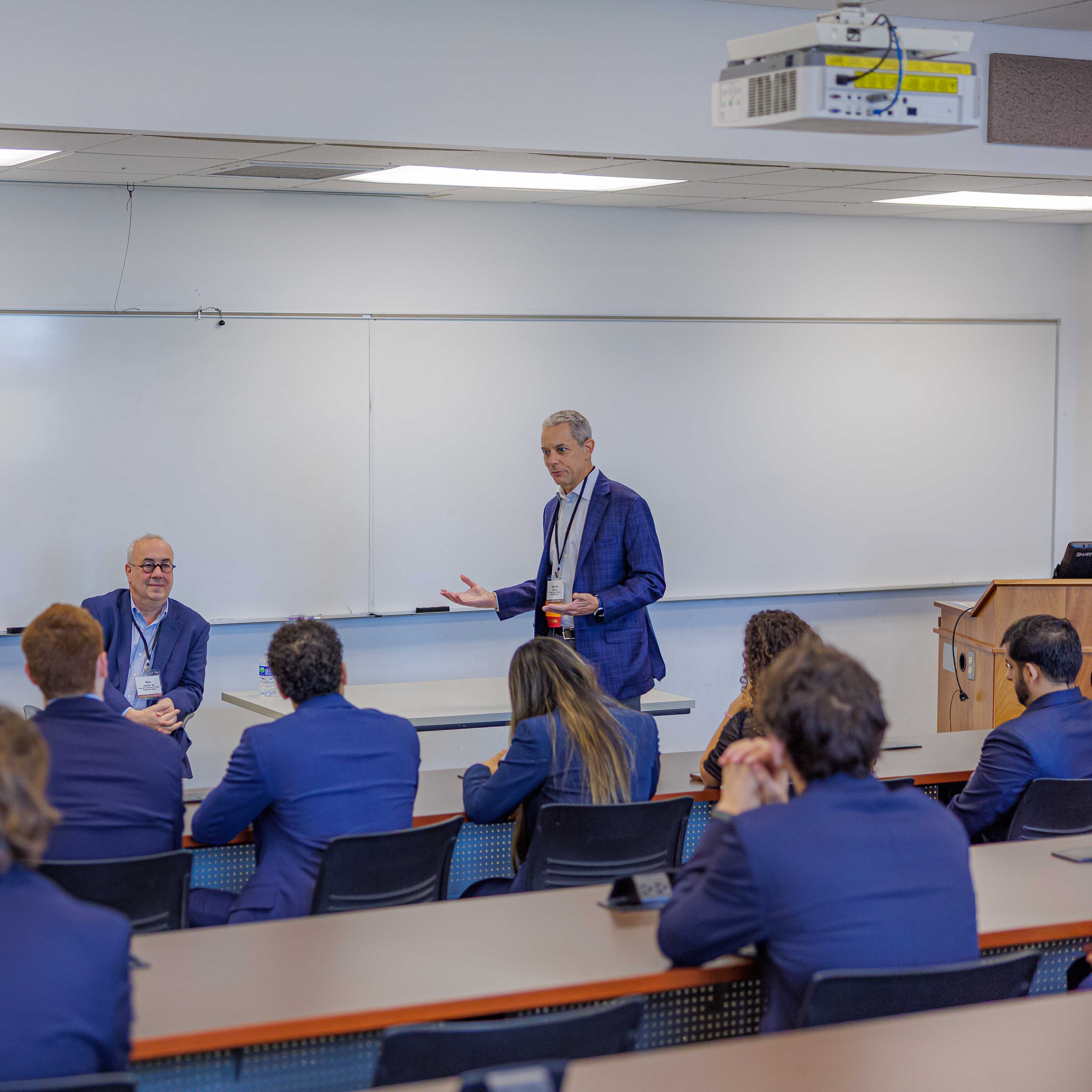 Andrew wears a blue suit and talks animatedly to a room full of attendees. The audience wears business attire.