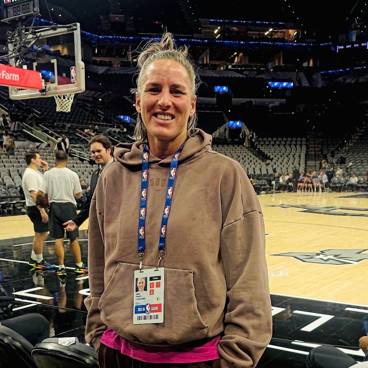 Anne Tierney stands courtside, smiling and wearing a brown hoodie with an id badge on a lanyard around her neck.