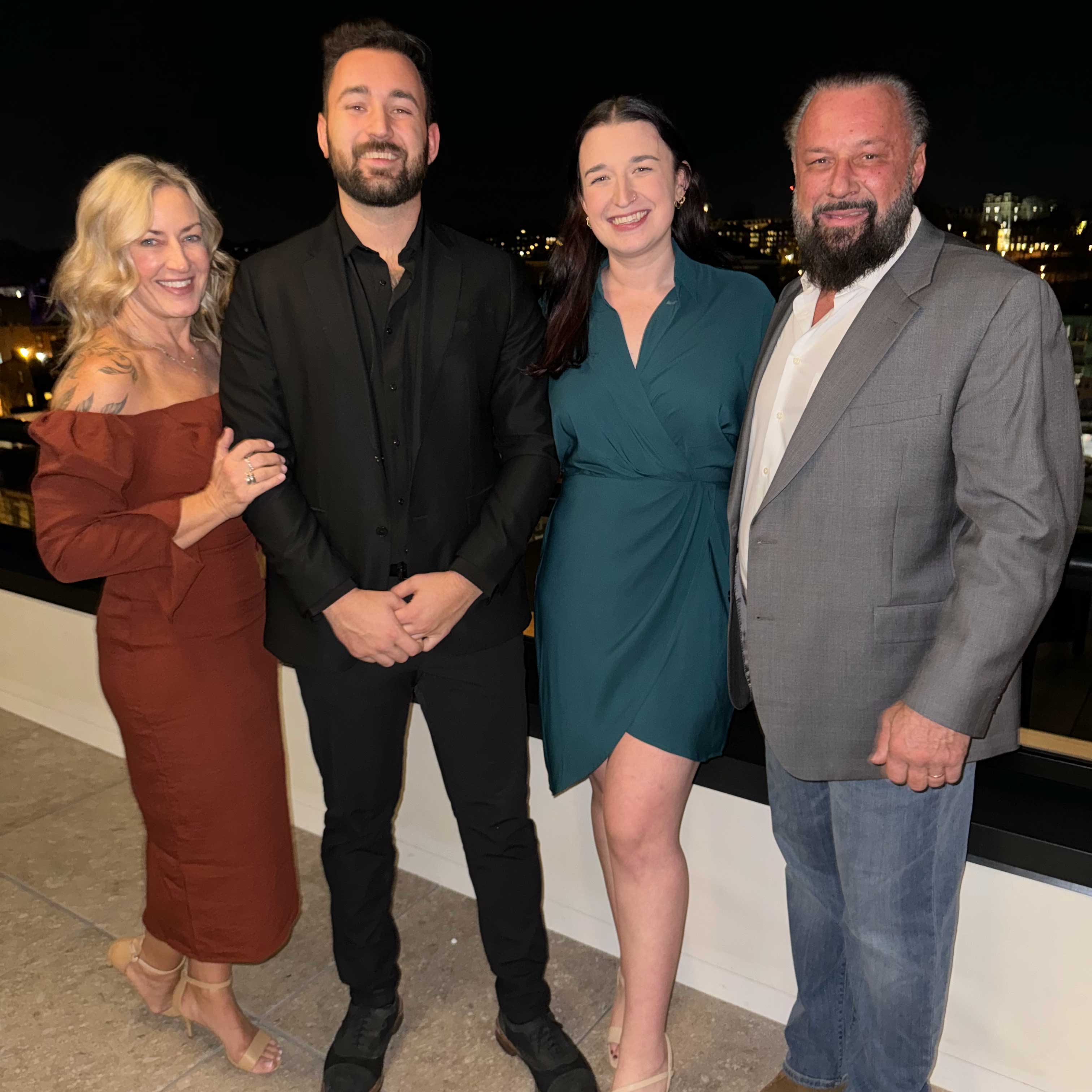 Patti, Anthony, Mara, and John Trapani stand on Zest's balcony at night. They wear business casual and smile. 