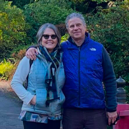 Wearing North Face Vests, couple stands on a walking path arm in arm and smiling.