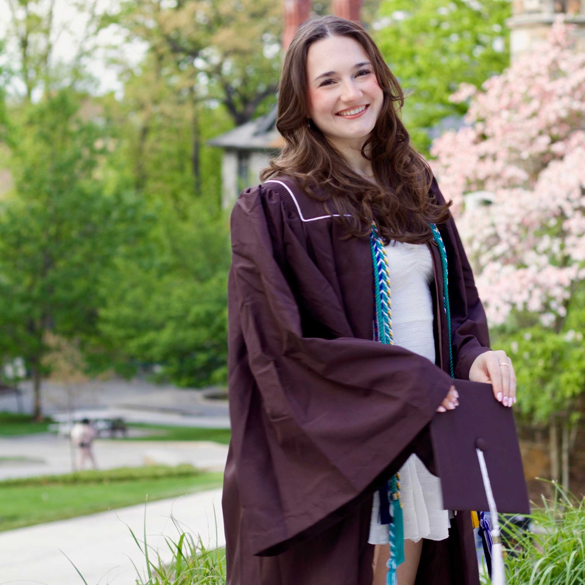 Ava wears her graduation gown and holds her cap while smiling on campus.