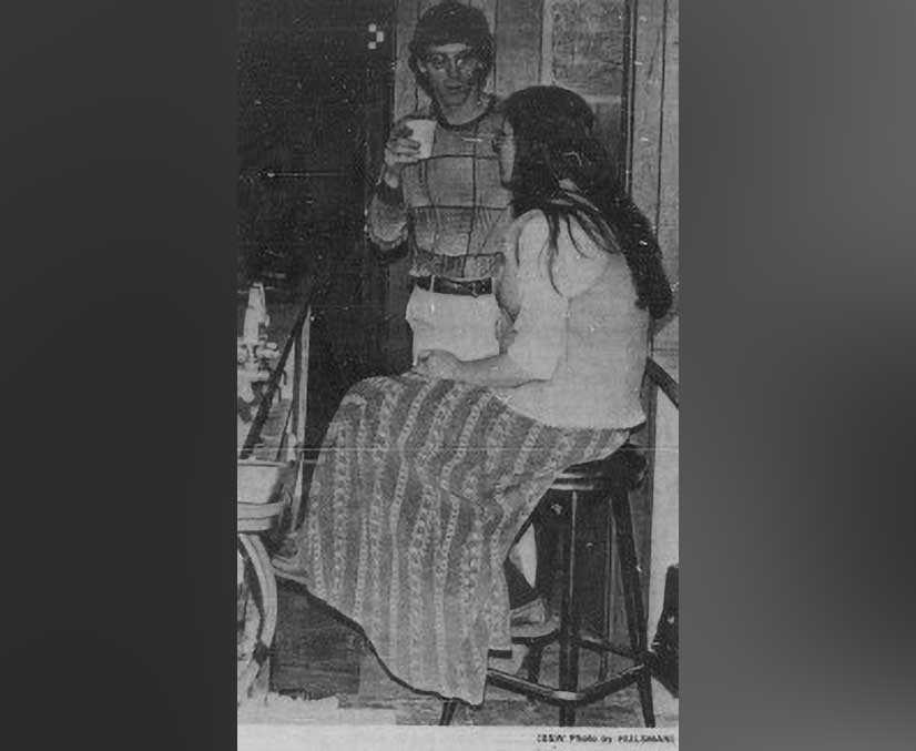 A black and white photo of two students chatting at a bar while one sits and the other holds a drink in their hand.
