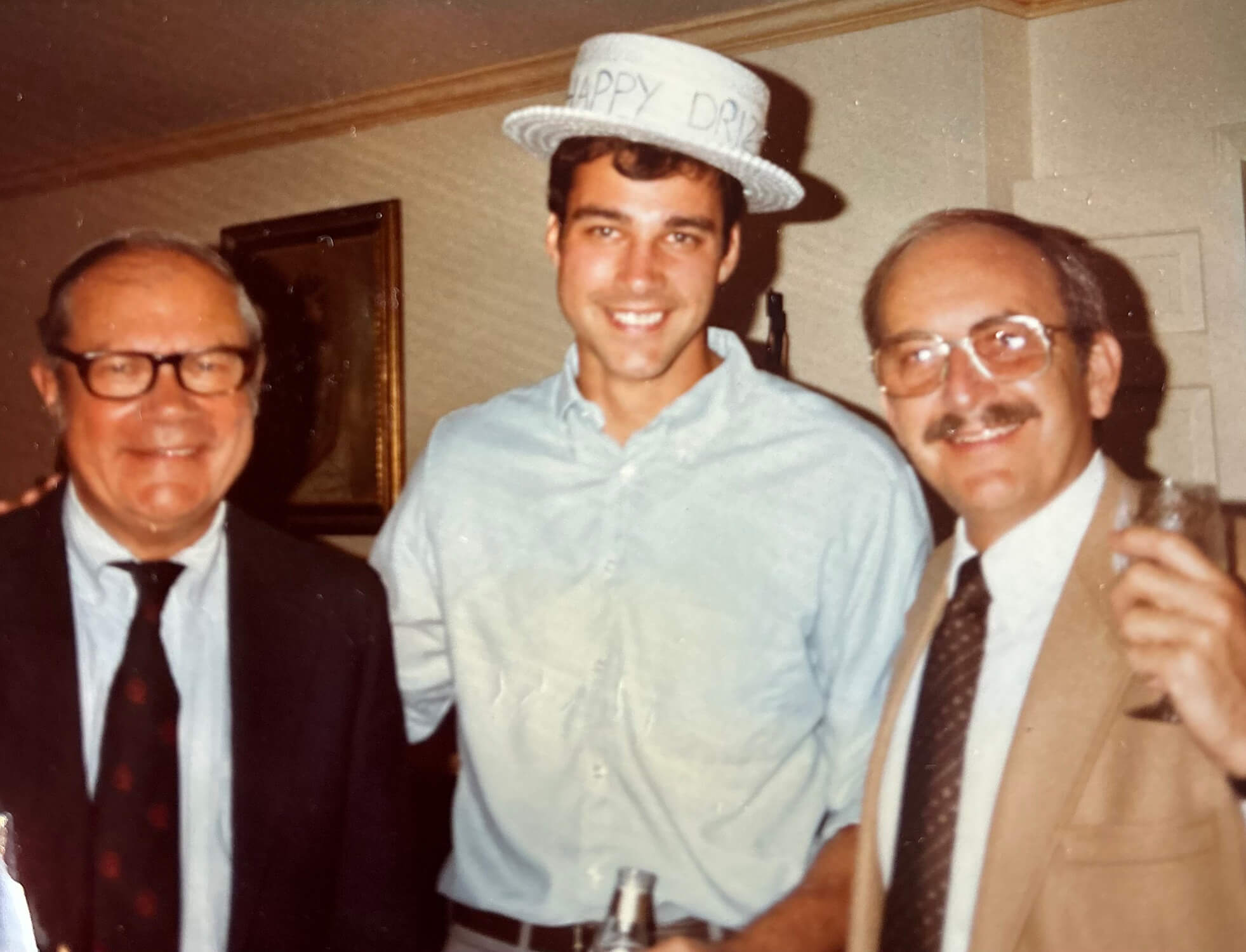 Tom Bartlett stands wearing a white boater hat with nondescript writing on the front with his father and father in law on either side of him.