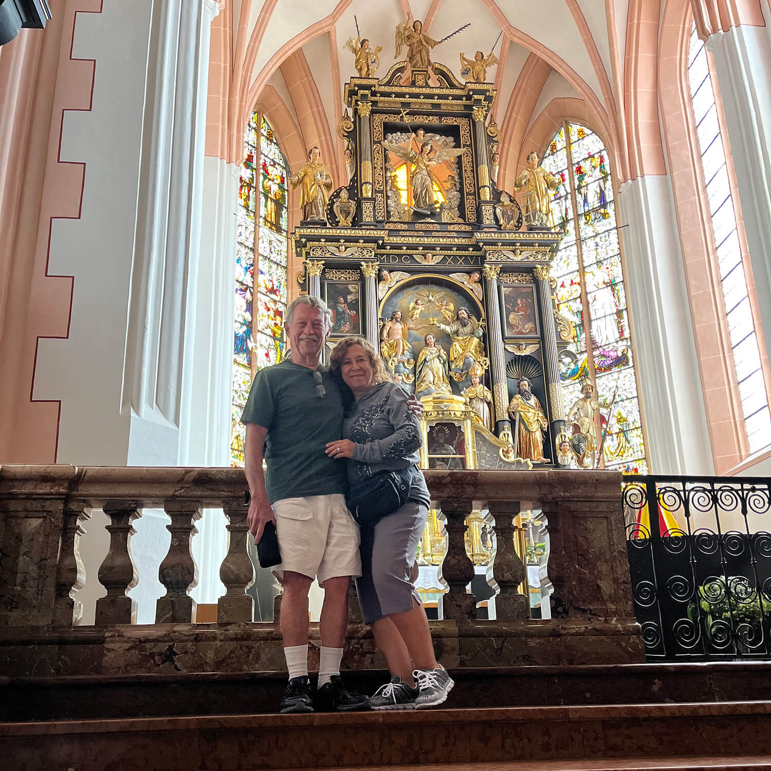  A husband and wife pose lovingly in each other’s arms in front of the alter of a chapel with ornate gold statues, stained glass, and icons behind them.