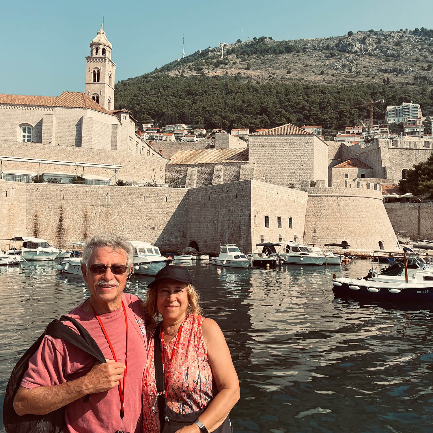 A man wearing sunglasses and carrying a black backpack on his shoulder stands with his arm around his wife in front of a canal with boats docked.