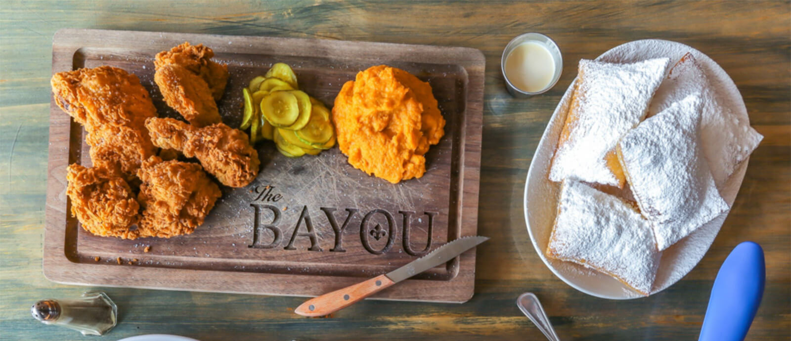 Fried chicken, pickles, a dollop of mashed sweet potatoes, and a small knife on a cutting board engraved with The Bayou.