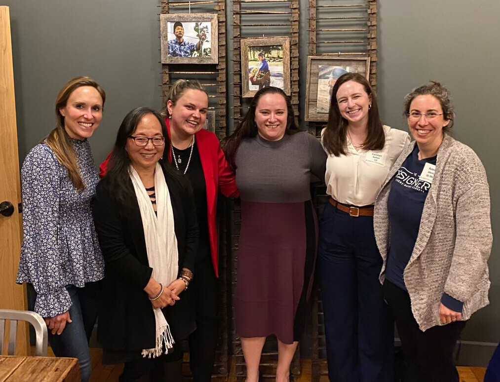 Six women stand side-by-side, arm-in-arm while smiling for a photo.