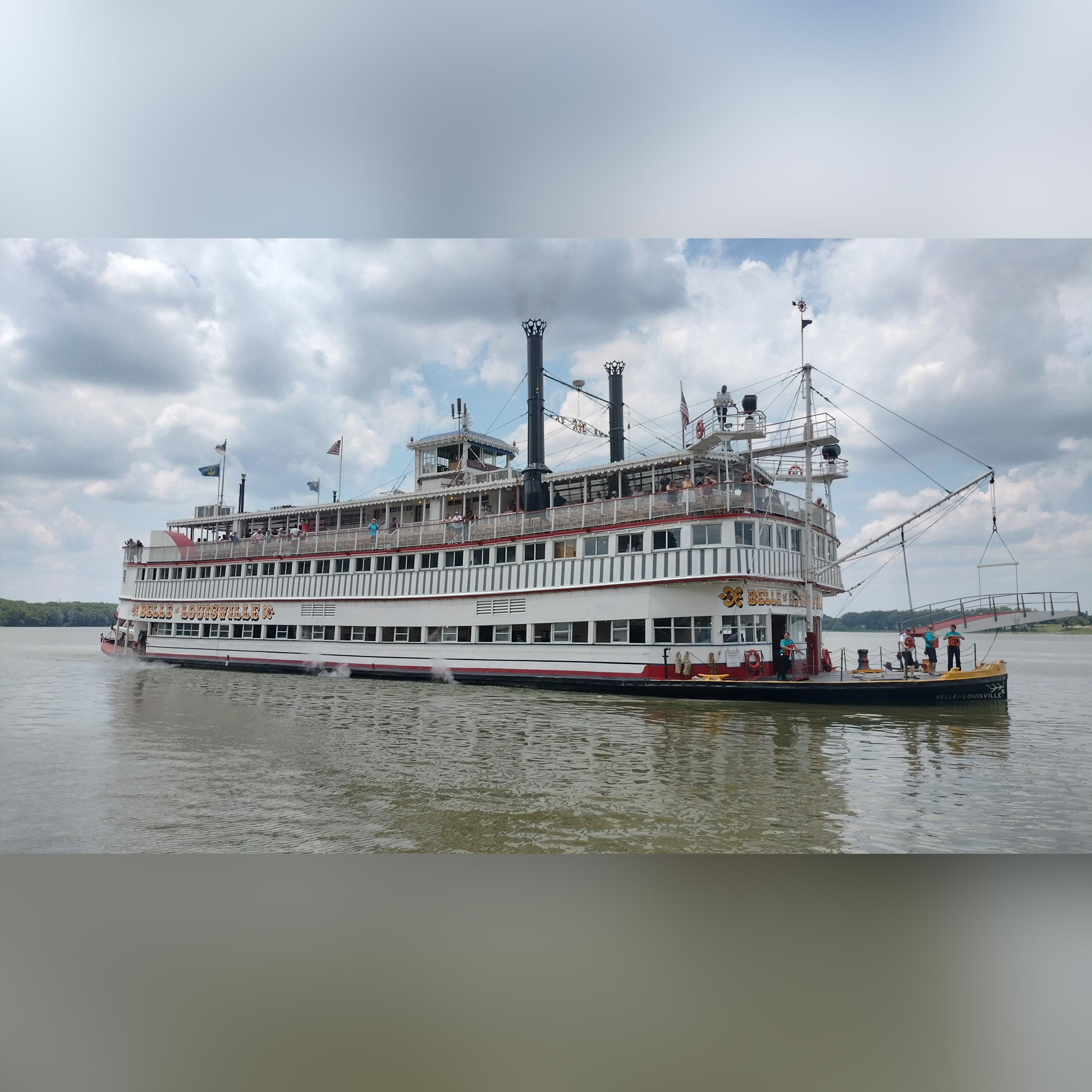 The Belle of Louisville cruises down the Ohio River on a cloudy day. People are visible on the decks.