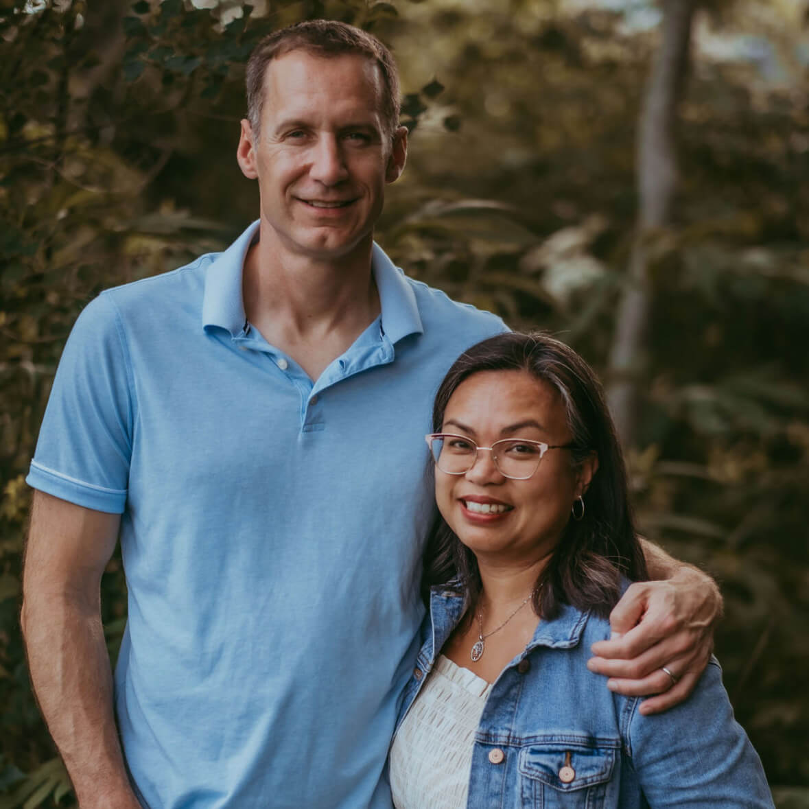 Tall husband wearing a blue polo shirt stands beside his wife wearing a denim jacket and glasses with his arm around her