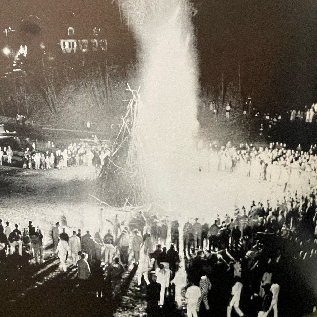 A black and white photo of a tall bonfire surrounded by a wide circle of people during the night.