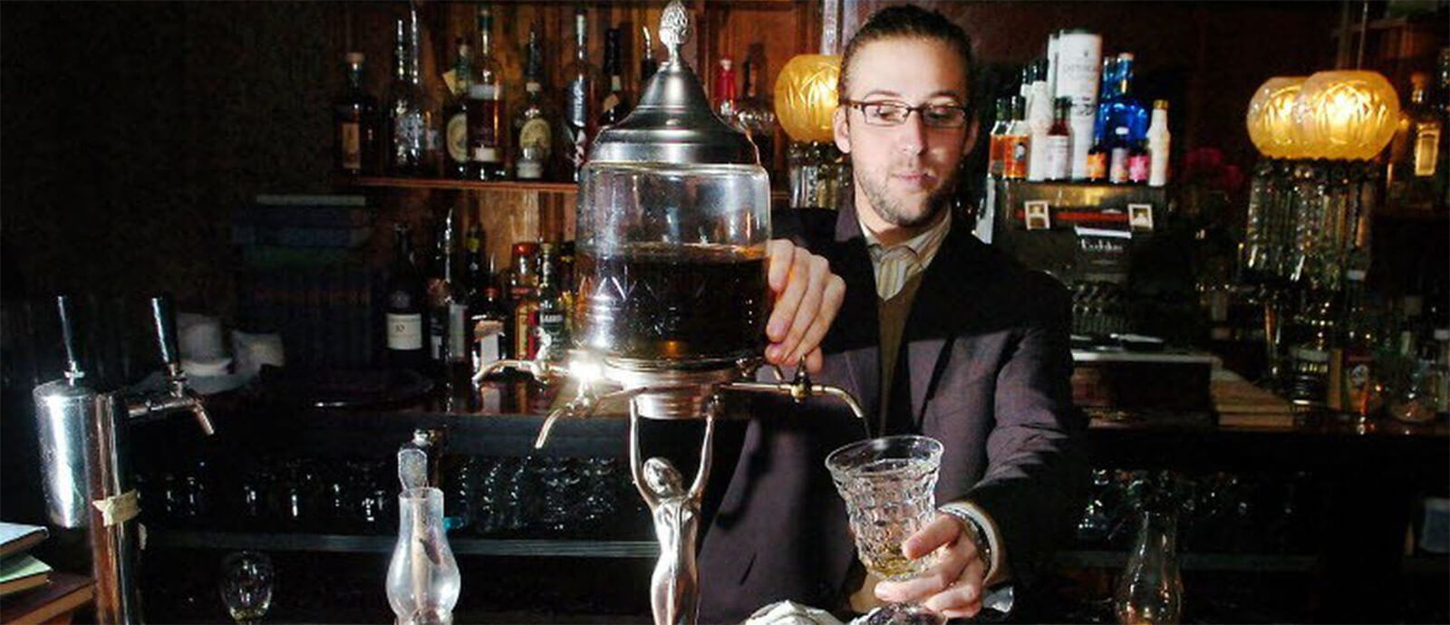 A man in a black suit jacket pours a drink into a crystal glass from an ornate liquor dispenser in front of a fully stocked bar.