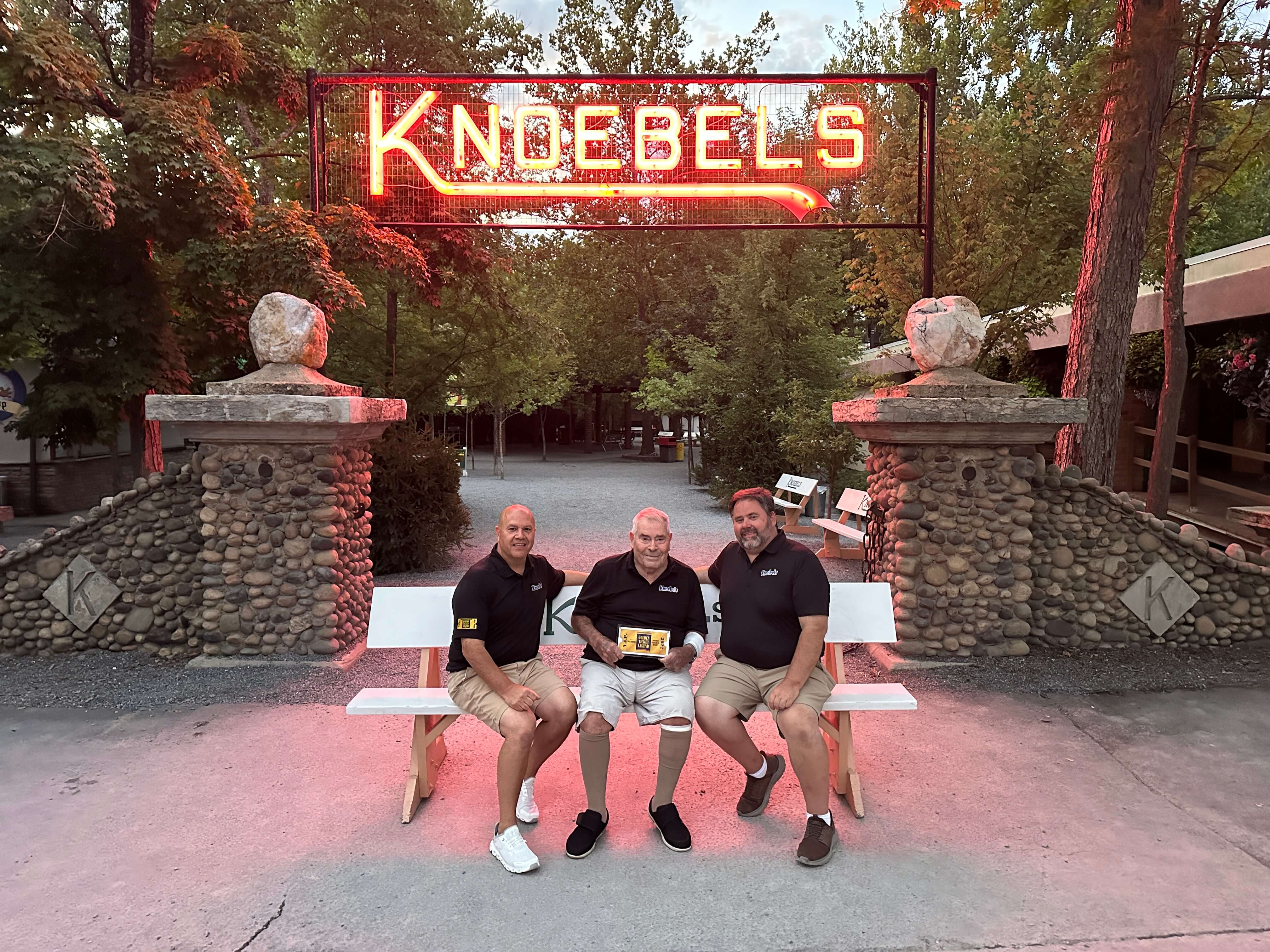 At twilight under a red neon sign that reads “Knoebels”, Brian and Rick Knoebel sit on a bench on either side of their father, Dick.