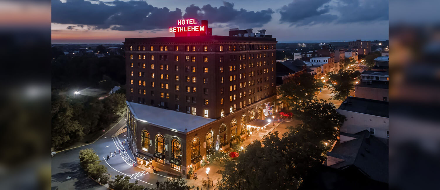 A large brick building with a bright red “Hotel Bethlehem” sign lit up against the evening sky.