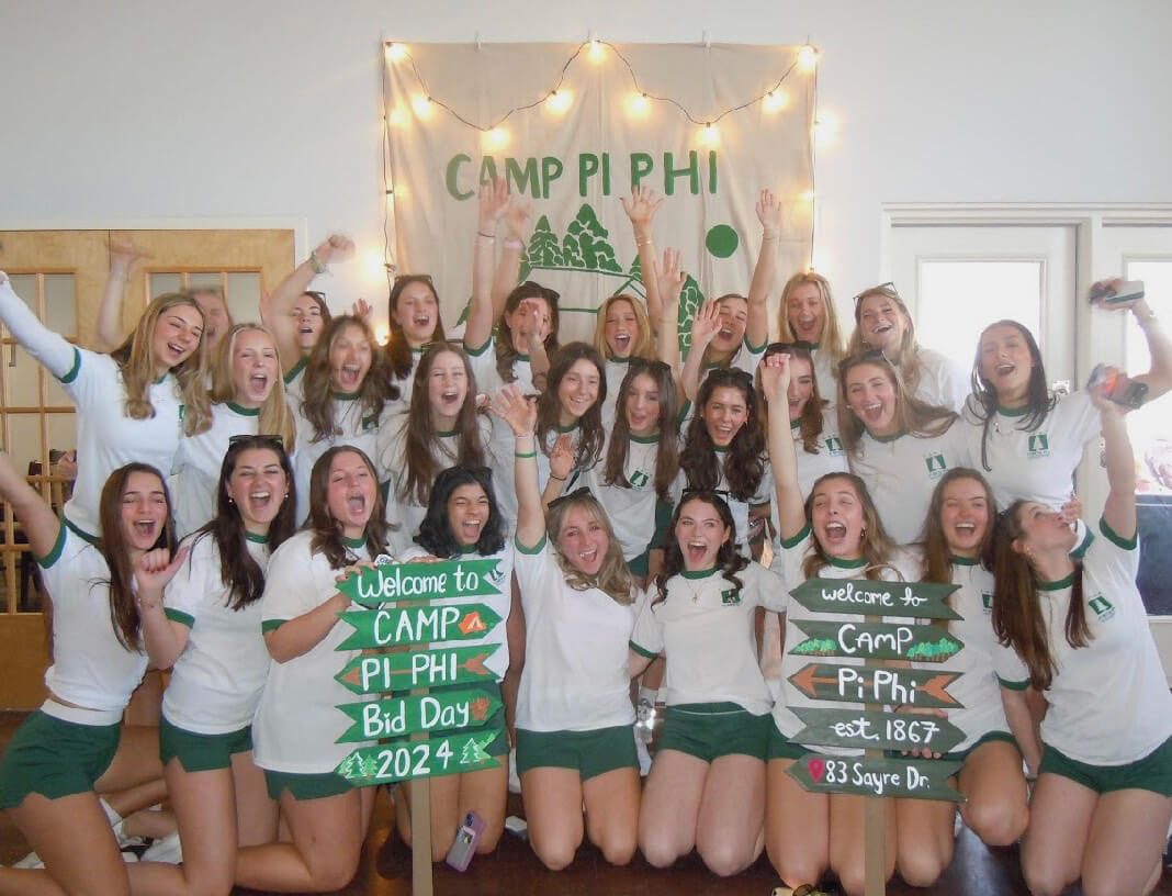 Wearing green trimmed white tshirts and holding camp-themed signs, students pose in front of a sign that says “Camp Pi Phi.”