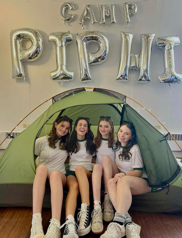 Four students pose in a camp tent, wearing green trimmed white tshirts and sneakers under balloons that spell out “Camp Pi Phi.”