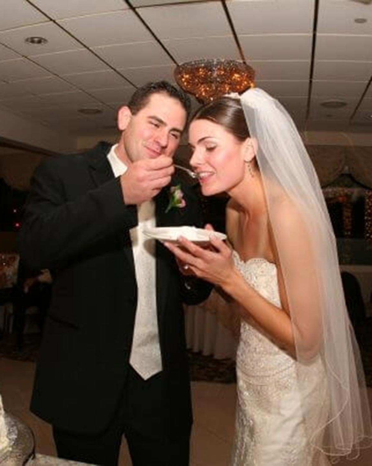 Kyle feeds Cara a bite of wedding cake during during their wedding; she wears a strapless gown and veil and he wears a black suit.