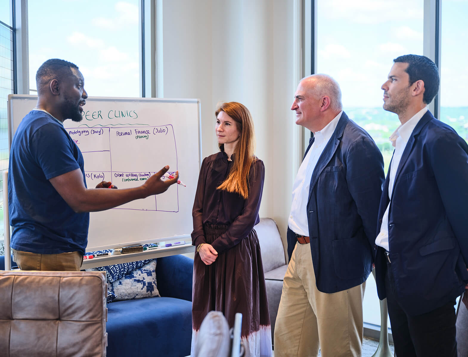 Three mentors stand listening to a man at a whiteboard that reads "Peer Clinics" with a marker in his hand and speaking.