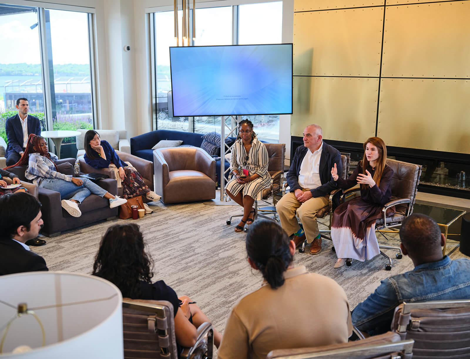 A group seated in a circle of comfortable chairs listens to a woman addressing the room motioning with her hands.