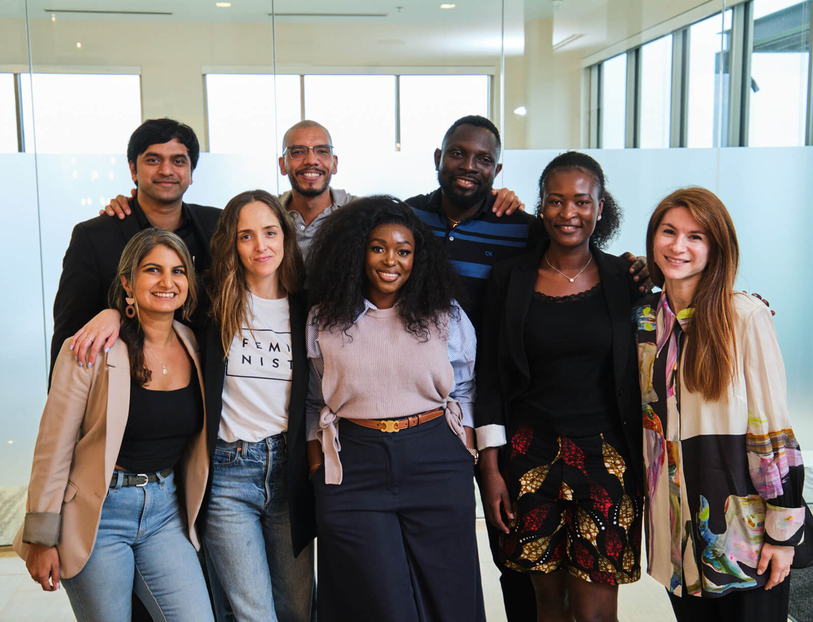 Eight people stand comfortably with arms around each other's shoulders, smiling at the camera.