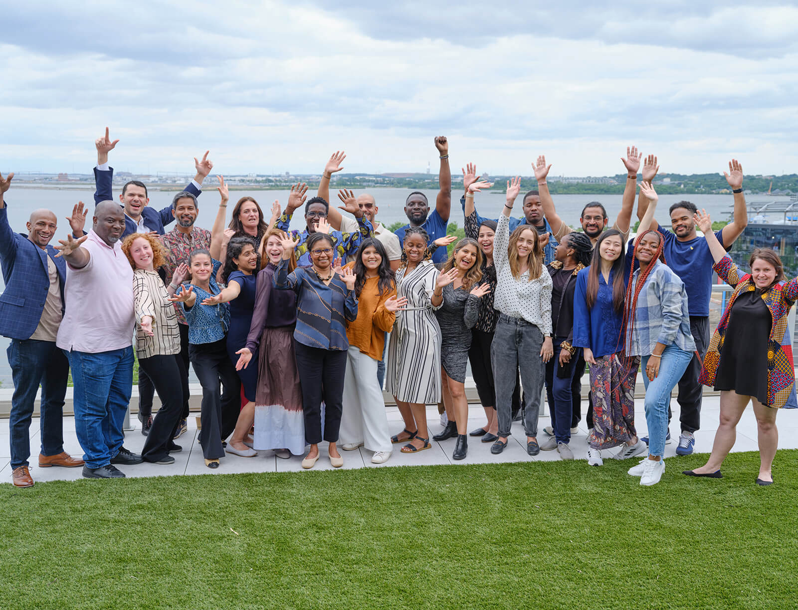 A large group poses for a photo along a river bank, raising their hands in the air enthusiastically.