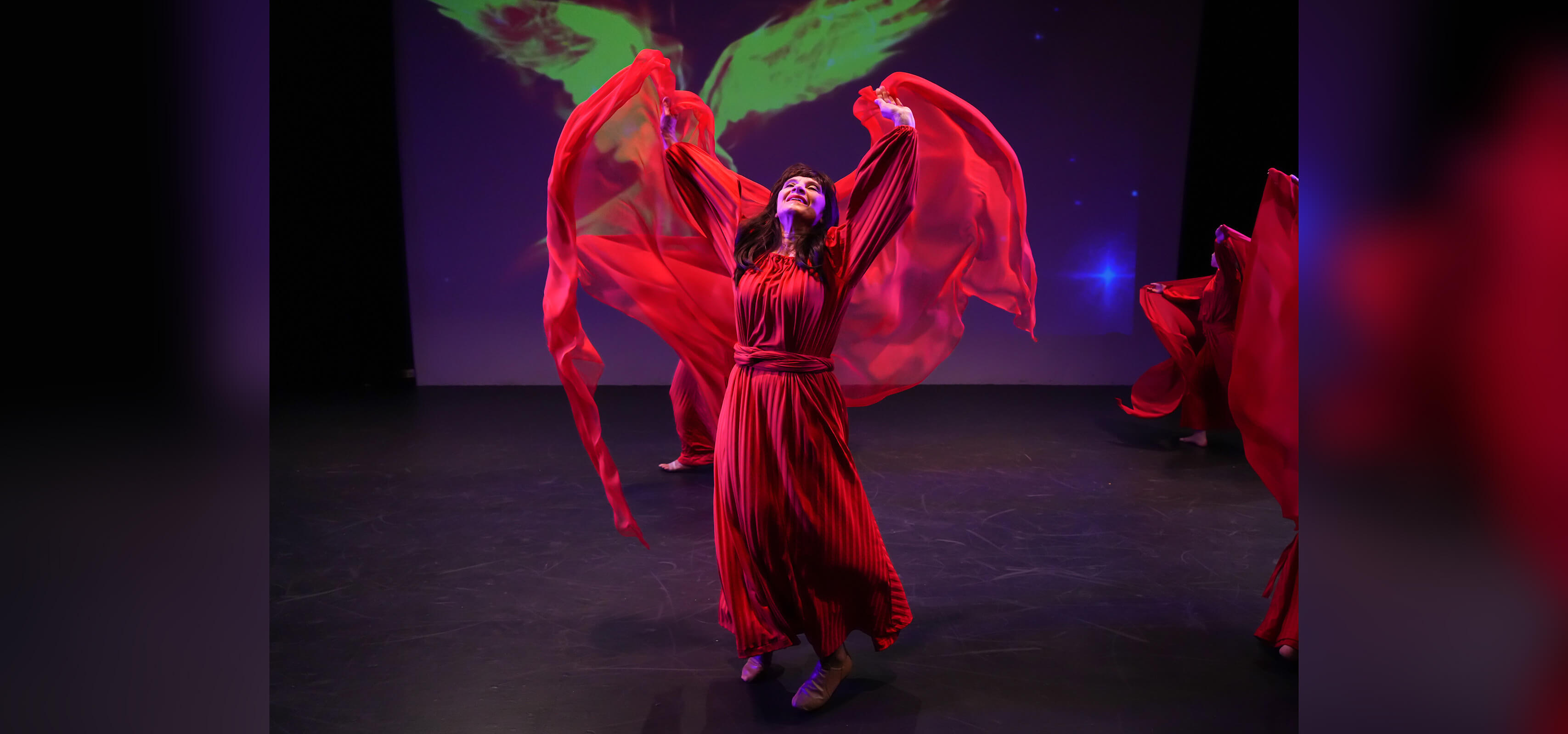 Female performer in a flowing red costume dances on a dark stage with sleeves floating in the air above her head.