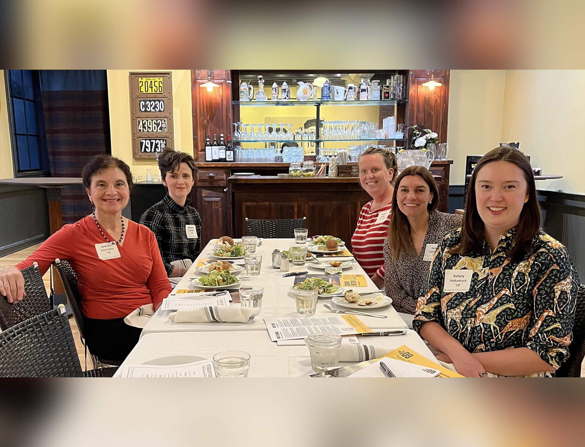 Salads sit in front of 5 women posing for a photo while sitting at a table.