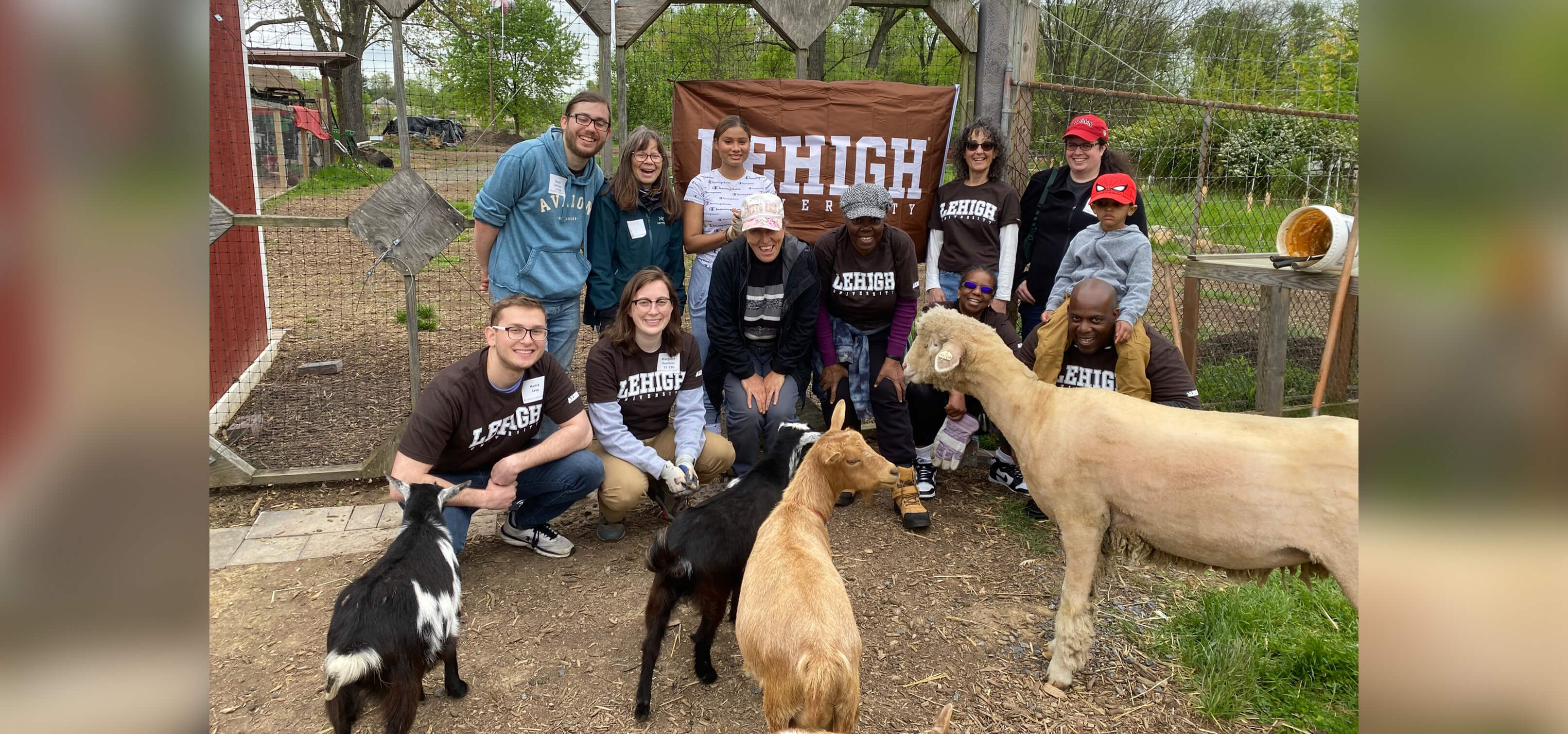 A group of alumni gather for a photo on a farm with goats with a Lehigh banner hanging on the fence behind them.