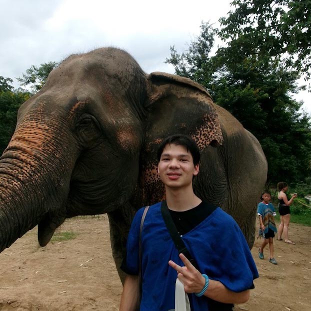Chancey stands in front of a large elephant in Thailand and smiles. 