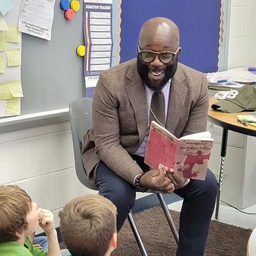 Charles sits in a classroom and reads to children. He wears a suit jacket, a tie, and slacks, and he smiles at them.