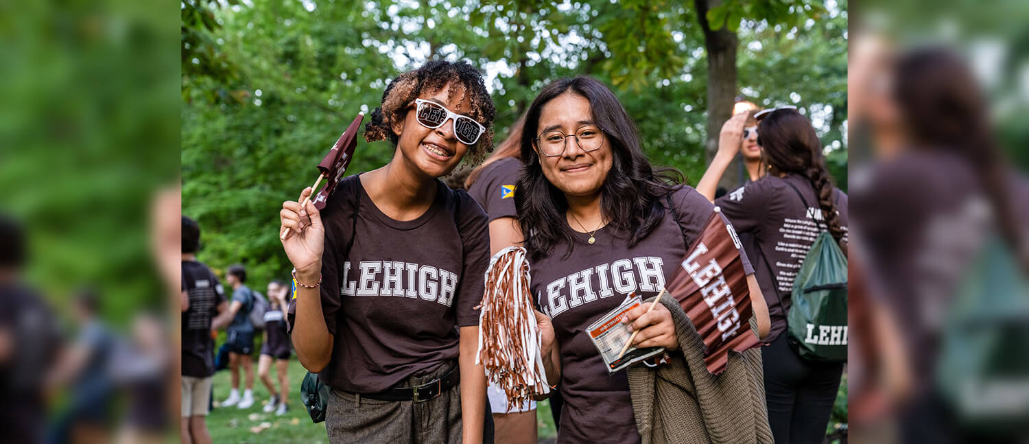 Two Lehigh students smile wearing Lehigh tshirts, Lehigh sunglasses, and holding Lehigh flags and pompoms.