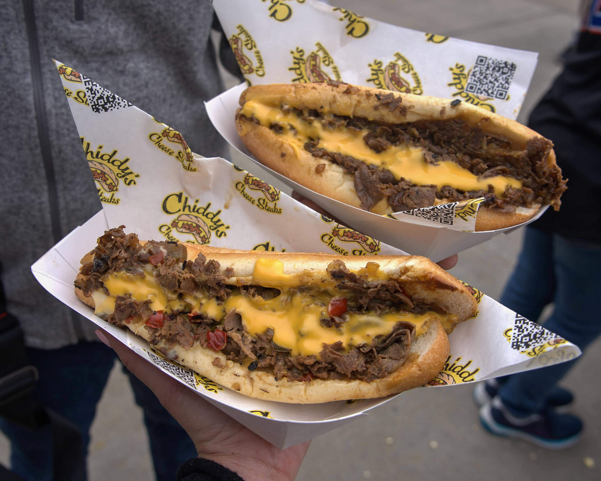 Hands holding two cheesesteaks on toasted long rolls, topped with Cheese Wiz, sitting on Chiddy's Cheesesteak wrapping paper.  