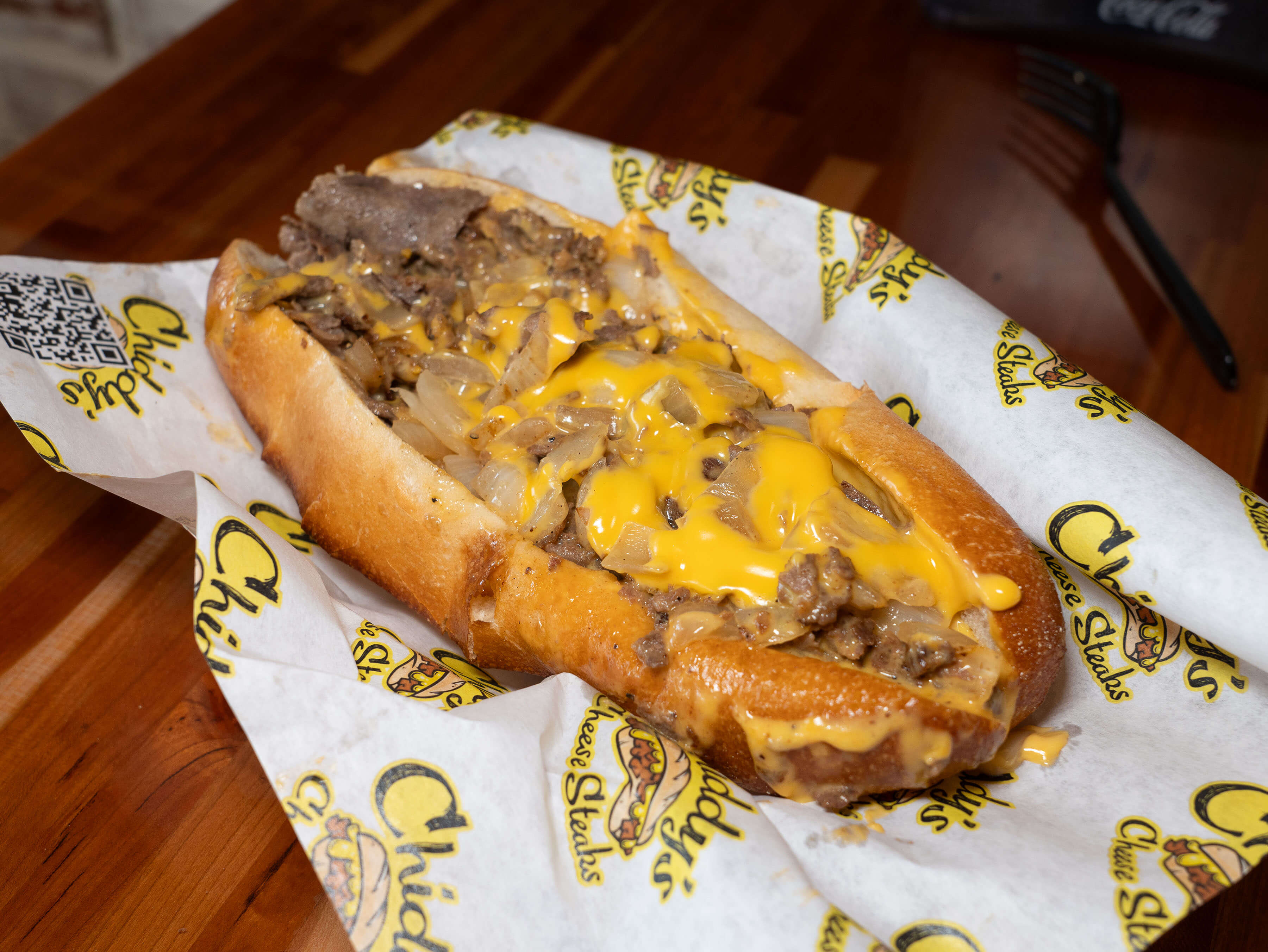 Wrapped in Chiddy's Cheesesteak wrapping paper, a cheesesteak on a toasted long roll is topped with fried onions and Cheese Wiz.  
