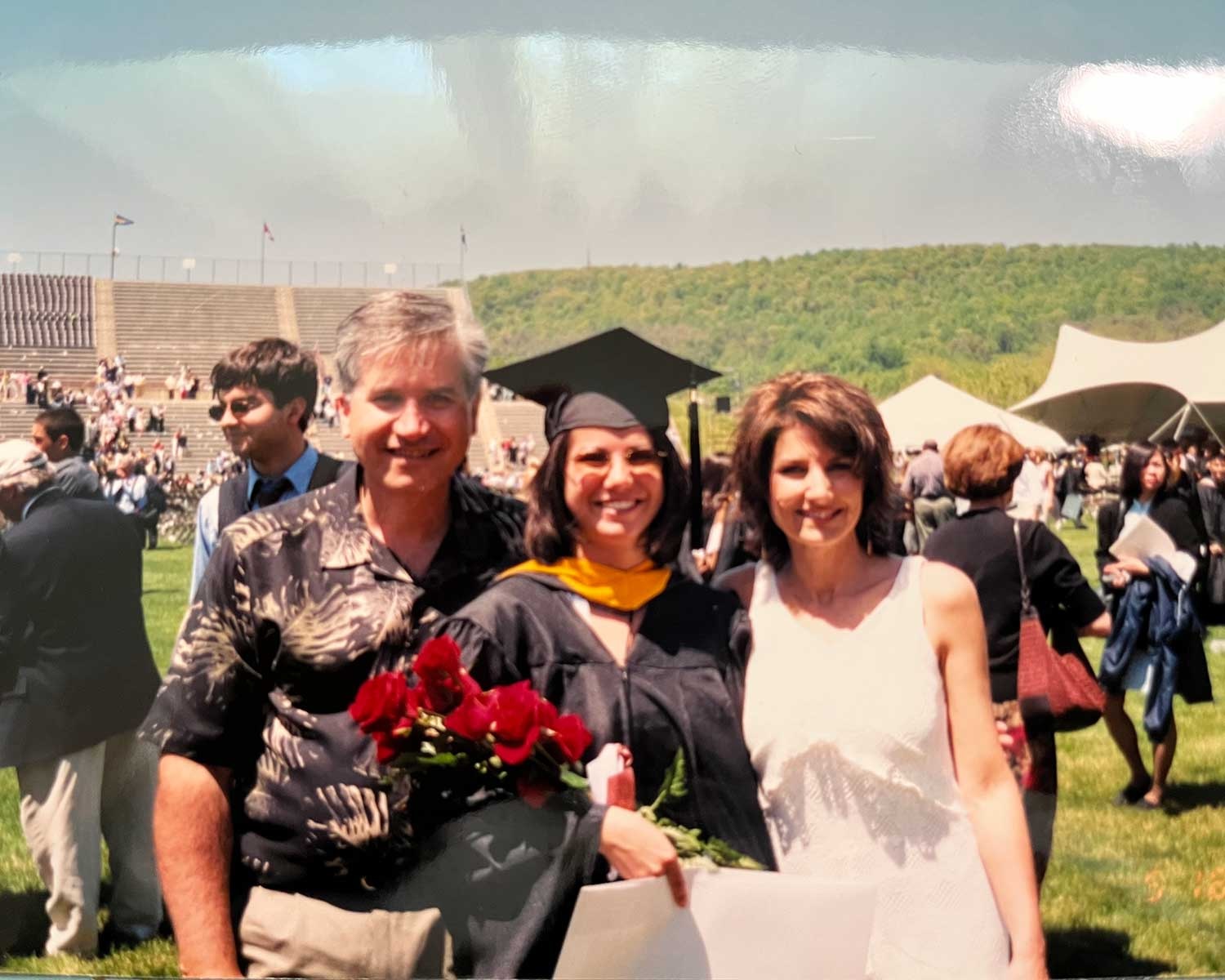 Christina stands between her parents at graduation. She wears full regalia and holds a bouquet of flowers.