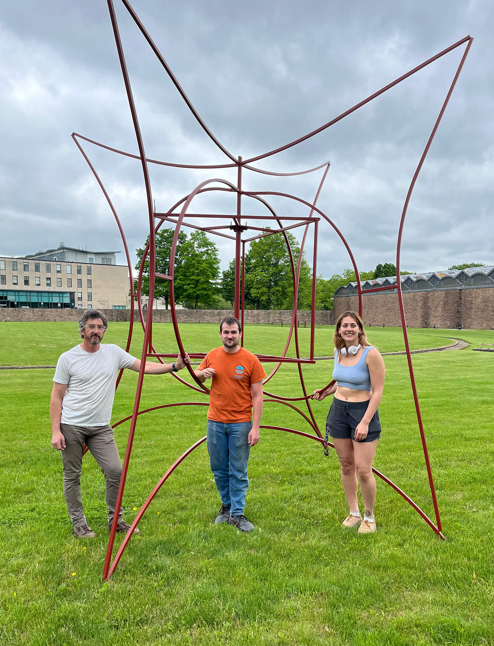 A large piece of red metal art stands in a green space with 3 contributors posing alongside it.
