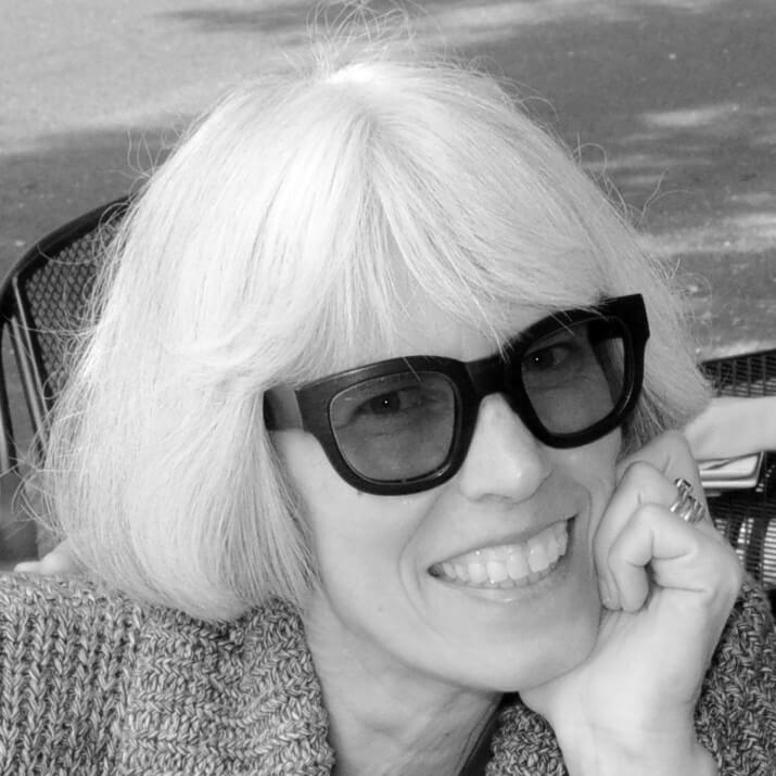 Black and white photo of Christine Ussler smiling in black framed sunglasses with her head resting on her hand.