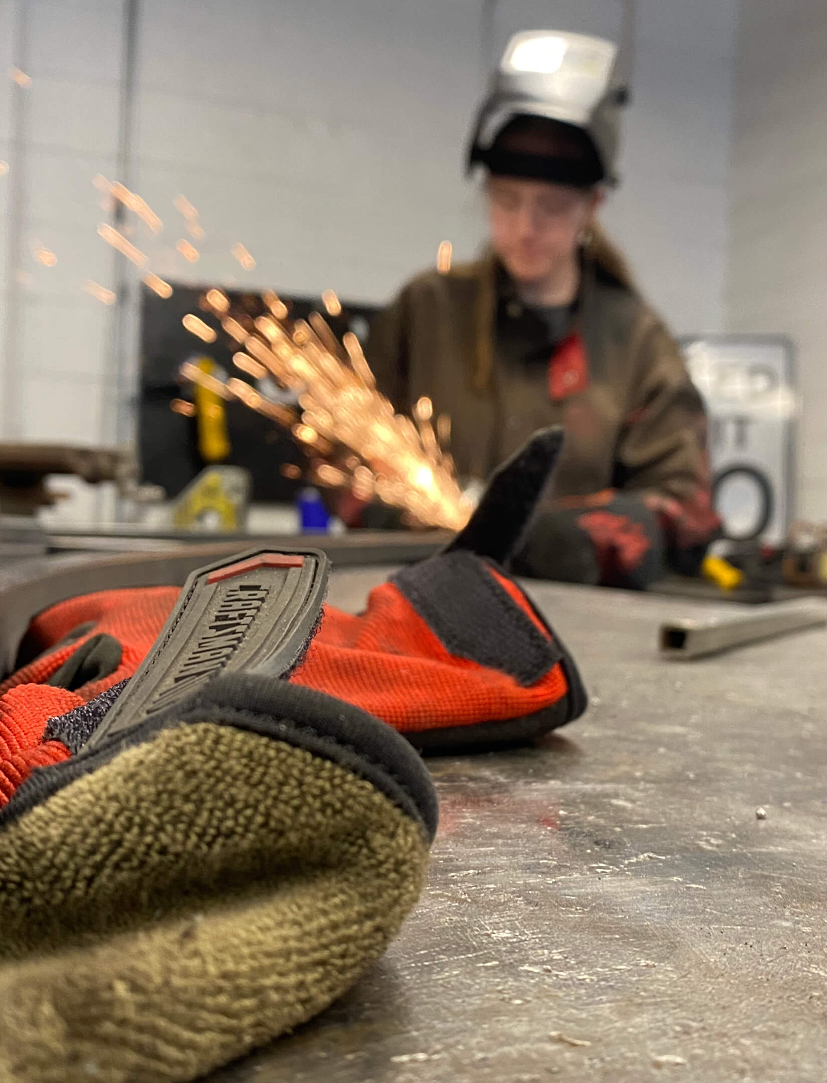 A welding glove sits in the foreground in focus while a student is out of focus in the background welding with sparks flying.