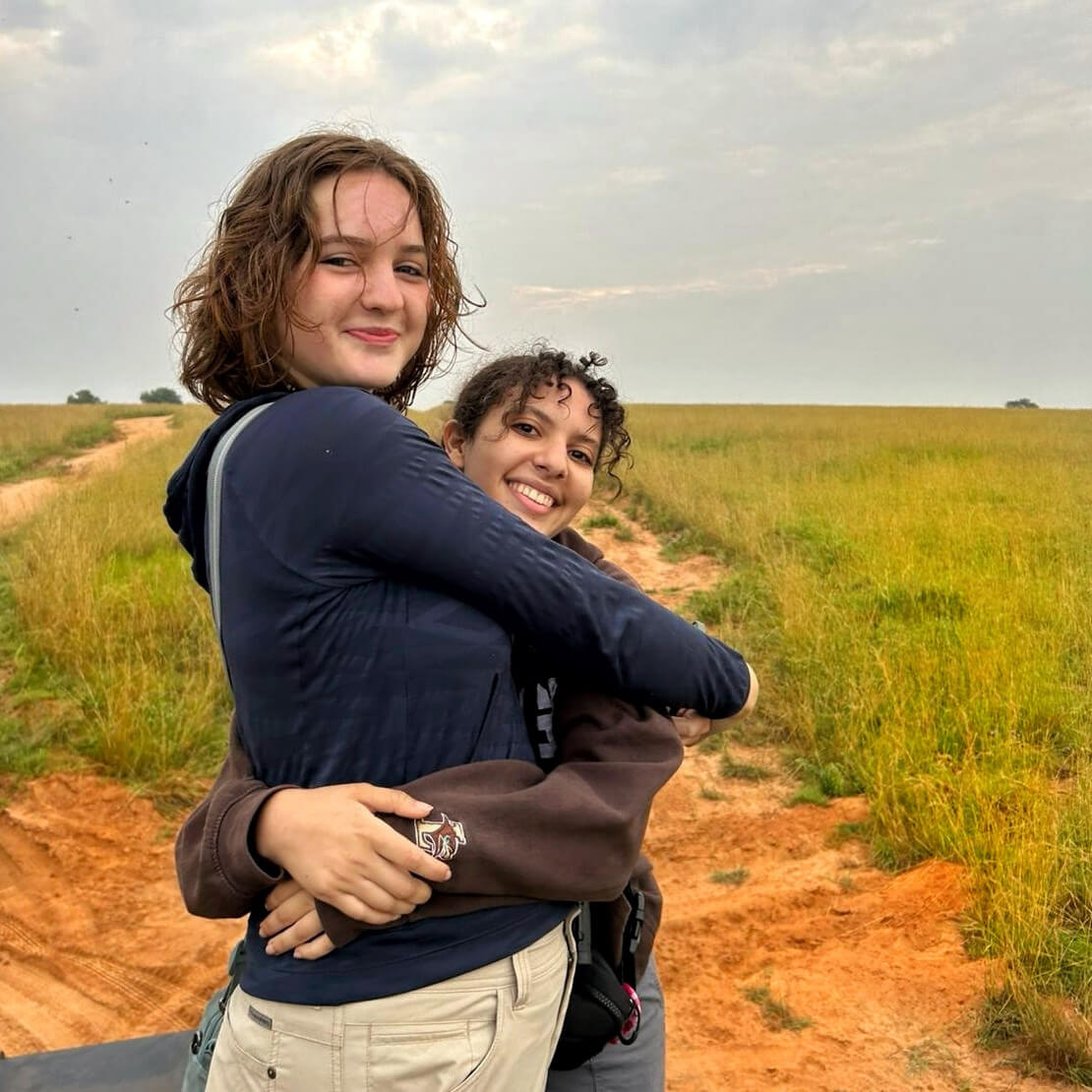 Standing on a dirt road in a field, Clara McAuley smiles closed mouthed while hugging a friend, also smiling for the camera.