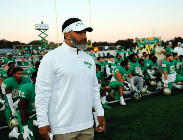 Coach Bryant Appling stands in front of his team on the sidelines while the knee behind him in green jerseys.